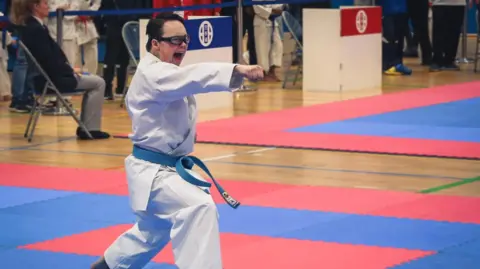 Family handout A teenage boy wearing a white karate outfit performs a punching move on a blue and red foam mat. He is wearing a blue karate belt, and glasses with a black frame. Behind him is a female judge sitting on a metal chair.