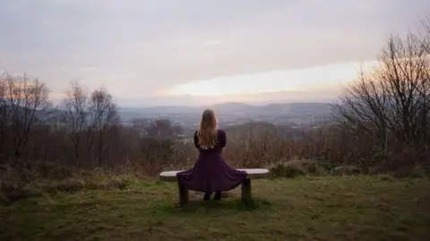 Getty Images A woman with blonde hair and a polka dot dress sits on a bench with a view of the skyline across the Malvern Hills.