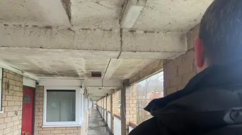 Simon Thake The back of a man's head as he looks down a corridor in a block of flats