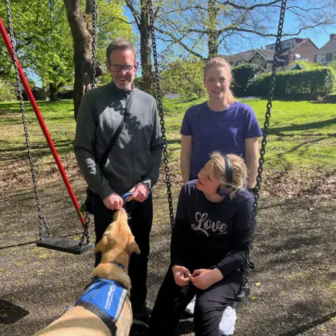 Rodney the support dog with Betsy at swings in a park with parents Miles and Laura Charlton behind