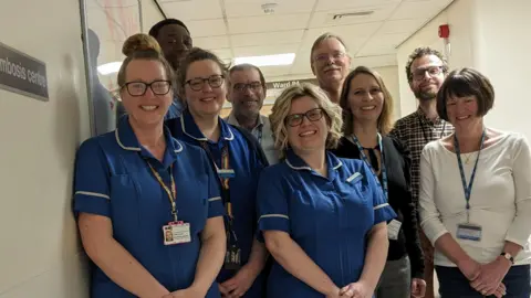 Sheffield Teaching Hospitals NHS Foundation Trust Nine people, some wearing blue scrubs, smile at the camera. They all stand in the corridor and look at the camera. Cathy Harrison is in public.