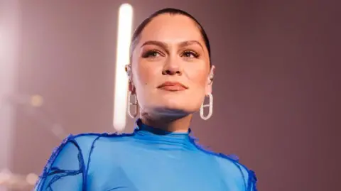 Jessie J wearing a blue lycra stage costume and large diamond earrings, smiling slightly on stage