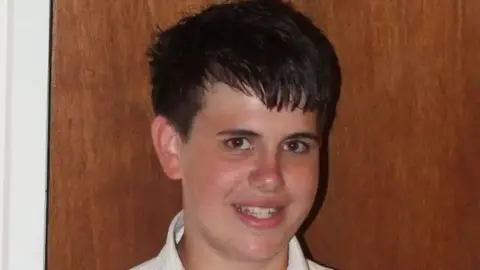 Jimmy Mizen smiling. It is a close up photo. He has dark hair and appears to be standing in front of an oak coloured door