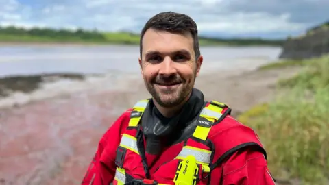A river rescue worker wearing red and yellow safety uniform. He has short dark hair and facial hair and is standing on the banks of a river.