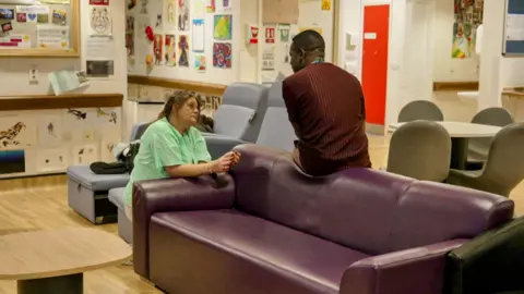 One man sat on a purple sofa, talking to a woman kneeling nearby, wearing a green hospital gown. There is also other seating there, a table and chairs behind the sofa.