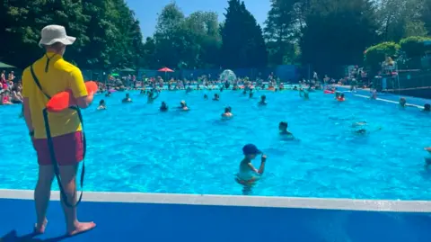 A man with a white hat, yellow tshirt and red shorts holding a long orange object looking across a blue outdoor pool with lots of people in it