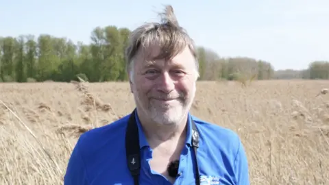 John Fairhall/BBC Dave Rogers stands in a wheat field and smiles at the camera. He has grey and brown hair, some of which rests over his forehead. He wears a blue top with a black binocular strap around his neck. 