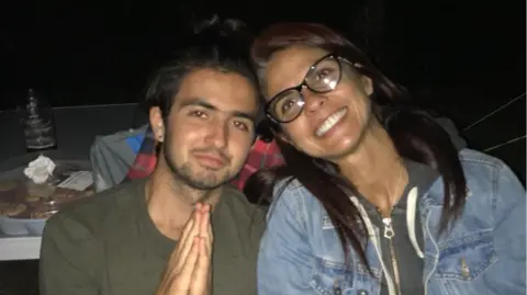 On the left of the image is Michael Frison, wearing a khaki green t-shirt. He has dark brown hair and some facial hair. His hands are placed together in a prayer pose. His mum, on the right, her head against Michaels. She has long dark brown hair, dark brown glasses and is wearing a denim jacket over a grey hoodie.