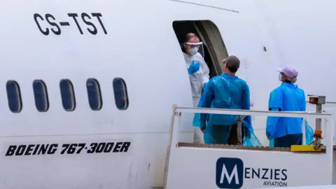 Man in full PPE talks to man and woman wearing blue plastic outer clothing and face masks at the entrance to a Menzies Aviation Boeing 767 plane.
