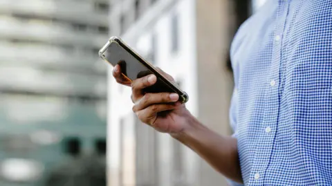 Getty Images A man in a blue and white checked shirt scrolls on his smartphone, with blurred buildings displayed in the background.