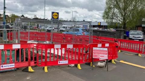 Temporary red construction fencing and silver construction fencing surround a damaged bridge in a town centre. There is a Lidl in the background, which has cars parked outside it.