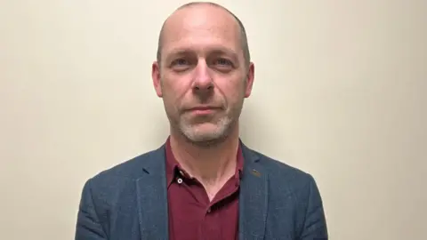 A man in a red shirt and blue jacket looking at the camera