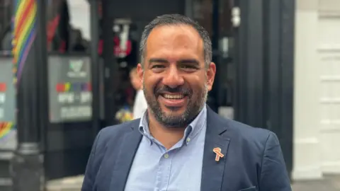 Stuart Antrobus/BBC Manuel Guerrero Avina staat op straat, glimlachend naar de camera, gekleed in een marineblauw jasje en een blauw shirt. Hij heeft een grijze baard en grijs haar.