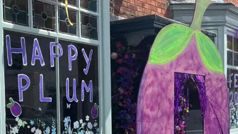 A window with "happy plum" written on it in purple, with plums also drawn on the window, and stickers of flowers and butterflies. A door is surrounded by a large painted cutout of a plum with streamers hanging in the doorway and flowers next to it.