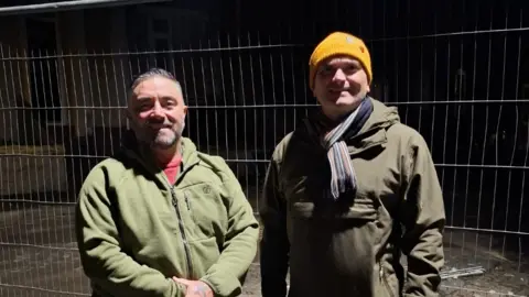 John Dunleavey standing with short hair and green jacket. Darren Parsley with yellow hat and green jacket and scarf. They are standing in front of a fence.