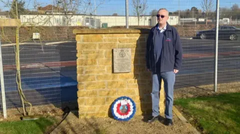 Geoff Bridgman is stood next to a wall with a plaque on it. There is a red, white and blue wreath on the floor, resting against the wall. Geoff has short grey hair and dark sunglasses on. He is wearing blue jeans, black shoes, a navy blue jumper and a navy blue coat with a logo on the chest.