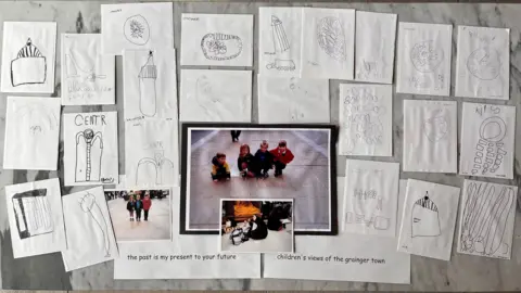 Newcastle City Council Various bits of white paper with drawings of buildings on them on a table. In the centre is a photograph of a group of school children crouched down on pavement.