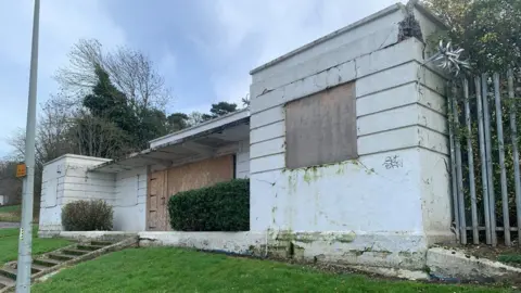 The entrance to the pool is boarded up and the white of the building has started to turn green and has cracks in places. 