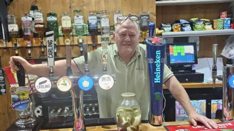 A smiling bartender stands behind a well‑stocked bar counter with multiple beer taps and shelves of spirits and snacks.