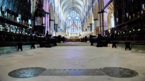 Lincoln Cathedral A dark piece of stone work on the floor of a cathedral with surrounding dark brown seats. The view looks across the choir towards a large stained glass window with intricate stonework.