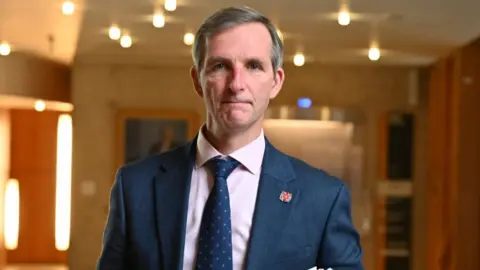 Liam McArthur, who has short greying hair, looks at the camera in a well lit room. He is wearing a blue suit and tie with a light pink shirt. 