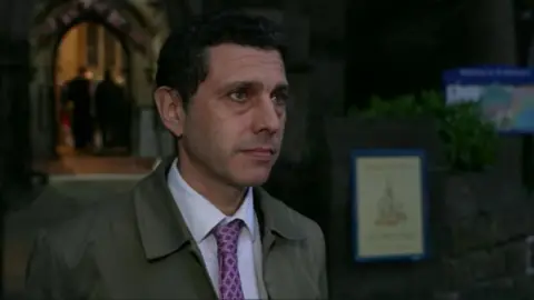 A black-haired man stands in front of the entrance to a church. He is wearing a grey coat, white shirt and a purple tie. A sign can be seen behind him to his right.