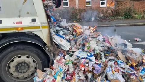 Piles of rubbish are tipped from the back of a bin lorry onto a road. There is smoke coming from some of it