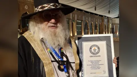 John Row John Row holding his Guiness World Record certificate for longest storytelling show. John has long white hair and a long white beard. He is wearing a black hat, a black jacket with gold details and a blue lanyard. He is also wearing glasses. He is looking at the camera and smiling. 