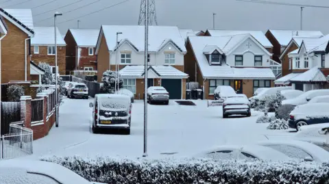 BBC weather watchers/Figaro Residential houses are blanketed with snow. The cars have a layer of snow on them, as do the roads and hedges.