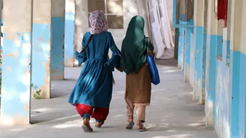 EPA Two Afghan girls walk down an outdoor hallway with blue and white walls, their hair covered. They wear bright colored clothes, and the sun is shining