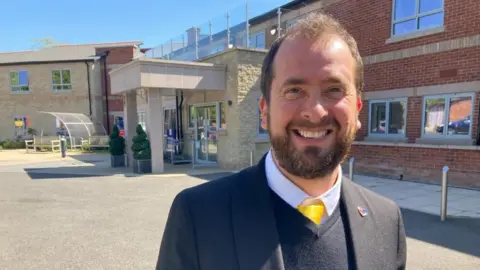 A man in a suit and tie is standing in front of a care home. He is smiling.