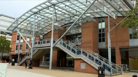 The outside of the site in Warwick Quadrant. It is a brown-bricked building with stairs on either side of the entrance and a glass roof