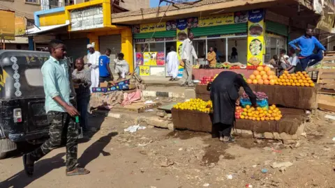 AFP via Getty Images A fruit vendor's wares are laid out in front of a partially ruined building. Someone is looking at the fruit and other people are walking around on the dusty ground. 