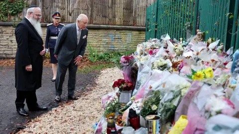 PA Media King Charles in a grey suit looks at flowers and tributes left outside the synagogue. Rabbi Daniel Walker is standing to his left.
