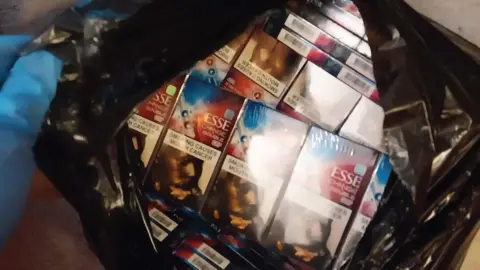 A gloved hand holds open a bin bag that is full of cigareetes