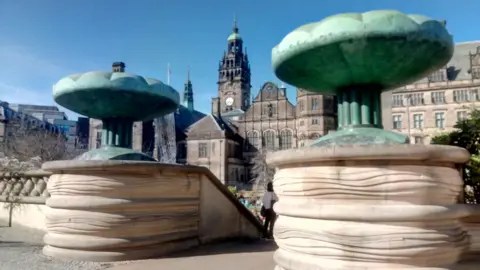 There are two large green stone structures at the top of steps leading to a paved area and fountains in Sheffield Peace Gardens. The town hall is in the background