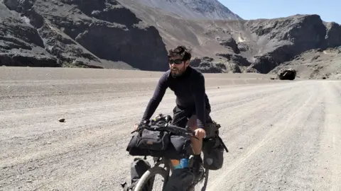 Nathan Hurley Man with dark hair and sunglasses cycles along a deserted track with mountains behind