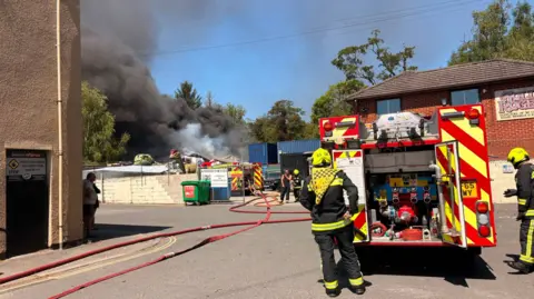 A fire engine with two firefighters dressed in uniform. In the background is another fire engine with a fighfighter speaking to a person dressed in black clothes. There is a road and two buildings. There are dinosaur statues and large plumes of grey smoke. The sky is blue.