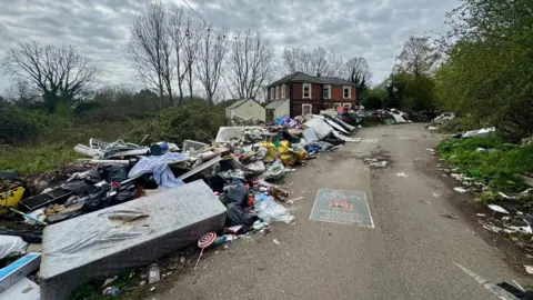 Fly-tipped rubbish piled up along a roadside leading to a red brick building and trees