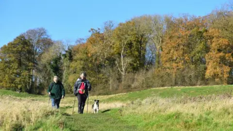 Lincolnshire County Council Two people can be seen in hiking gear with walking sticks walking through a field with a path in the middle. A medium-sized dog which is white and black is walking alongside them. There are trees in the background with a tinge of orange.