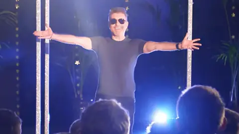 Simon Cowell surprises the pupils who are in a semi circle gathered around him laughing and cheering, some seen from the back