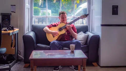 Eddy playing the guitar while sitting on a sofa next to a window, with a coffee table in front of him