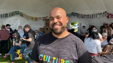 Tom Jackson/BBC Shaun smiles at the camera, standing inside a dome tent filled with people sitting around behind him. Colourful decorations hang from the sides. He has a cropped goatee beard and wears a grey T-shirt with the word CULTURE in bright colours.
