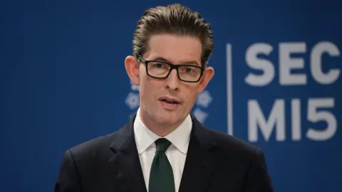 PA Media Sir Ken McCallum, a white man with dark, swept-back hair and rectangle, dark rimmed glasses, wearing a dark suit with a white shirt and dark tie, pictured in close-up with a long lens as he speaks. In the background behind him is the word MI5