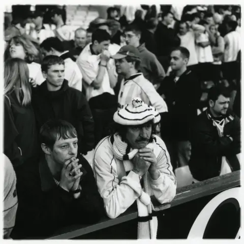 Jason Scott Tilley/Lower Block A black and white image of Coventry City fans leaning on a rail. One is wearing a white top, scarf and knitted hat. 