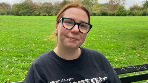Sofia Luis-Hobbs/BBC A woman sitting on a park bench. She is wearing a black t-shirt with white writing. She has glasses and ginger hair. In the background, there is grass and trees.