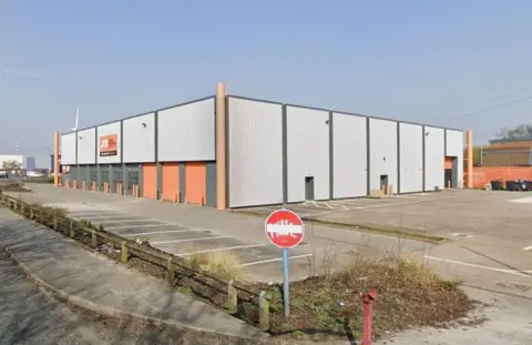 Google A pale grey warehouse photographed from the road. It looks empty, and there is graffiti on a road sign at the entrance to its car park. It has orange signage on it, and three orange garage doors along one of its sides. There is another garage door on another side of the building which looks to be open, and there are industrial bins in its car park. The grass borders are messy and unkempt.