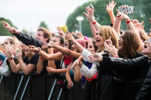 A crowd of people hanging over a barrier at a concert with their hands in the air, singing along. The crowd is mainly made up of young people with different coloured clothing on and different hairstyles.