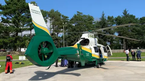 The Great North Air Ambulance Service helicopter from the tail end, on a sunny day, with emergency workers standing on grass at either side.