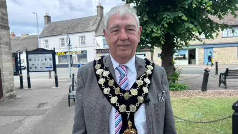 Kate Bradbrook/BBC A suited older man wearing the golden chains of a mayor is stood near a green area with independent shops across the road behind him